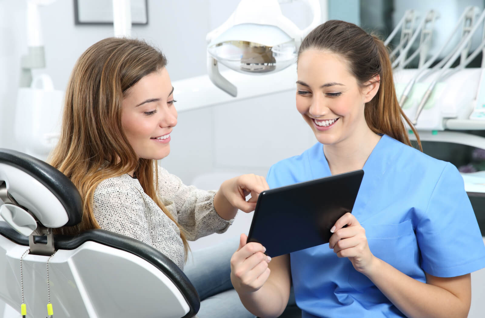 A female dentist in blue scrubs and a woman sitting in a dentist's chair looking at a tablet and discussing Invisalign.