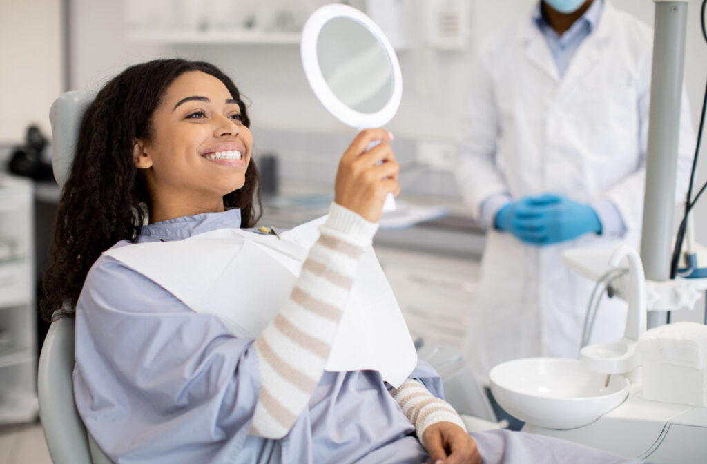 A woman sitting in a dentist's chair, holding up a mirror and smiling with his dentist in the background.
