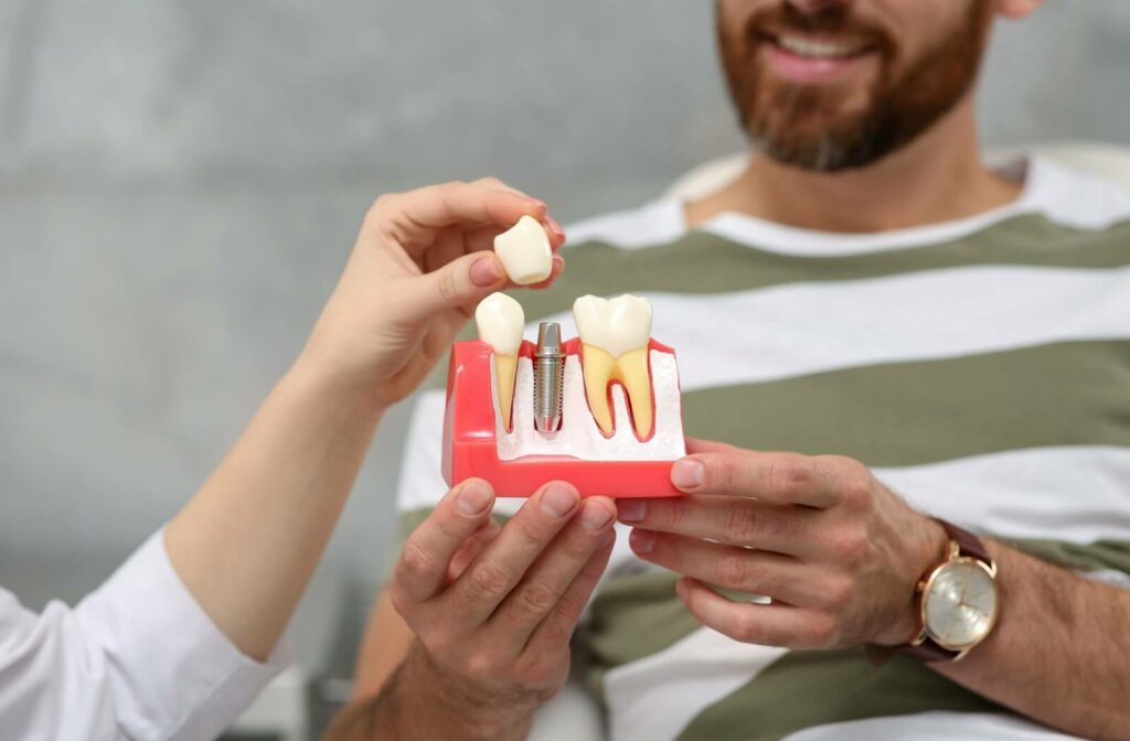 A close-up image of a patient holding a model showing a tooth implant and a dentist placing a crown over the post.