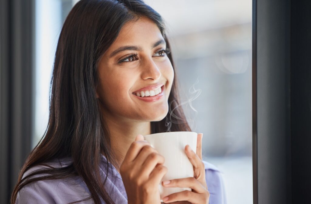 A woman smiling while holding a white coffee mug near a window.