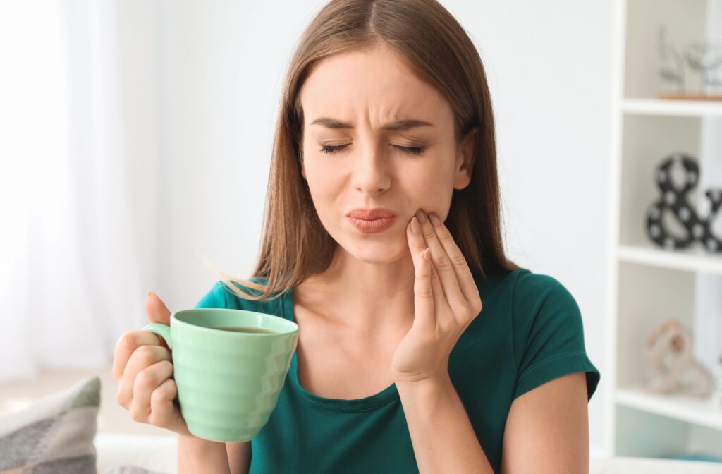 Woman with closed eyes holding her cheek in pain while drinking from a green cup, showing symptoms of tooth sensitivity.