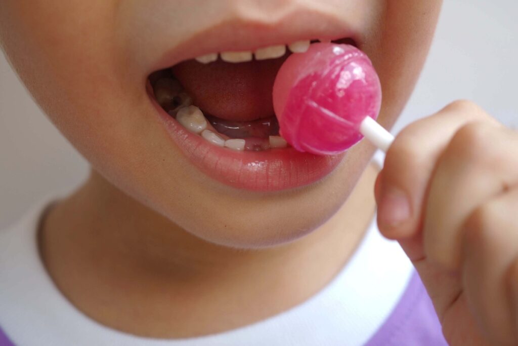 Close-up of a child eating a pink lollipop with visible cavities on their lower teeth.