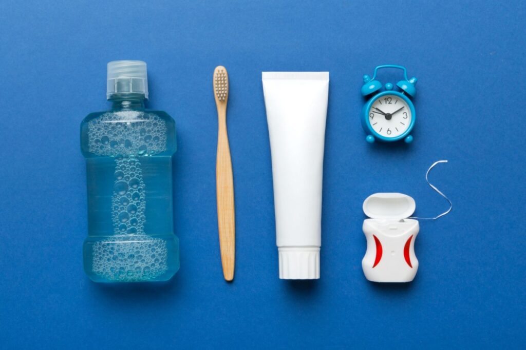 Dental care essentials including mouthwash, toothbrush, toothpaste, floss, and clock on blue surface.